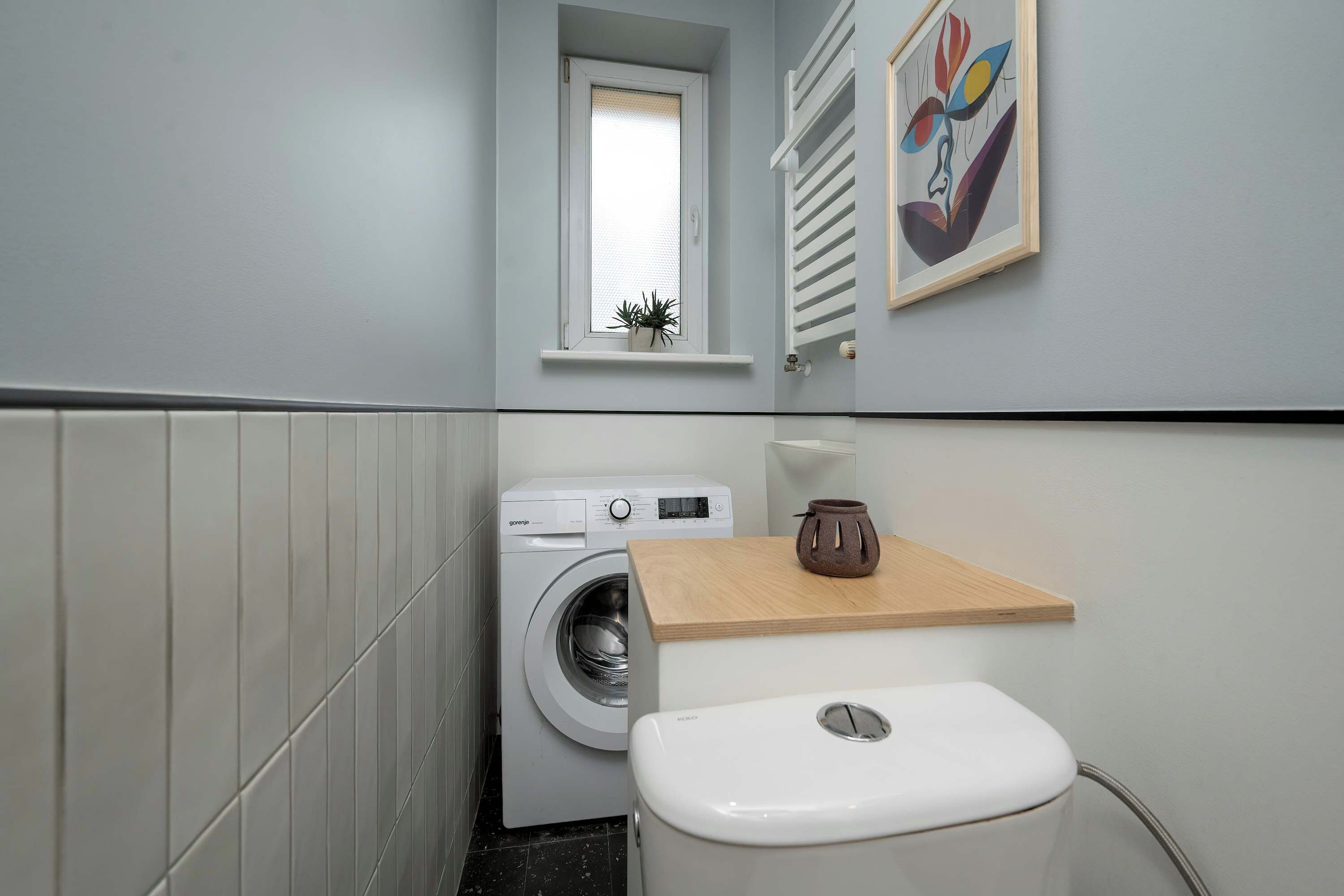 Een kleine wasruimte met een wasmachine, toilet en een klein raam met een plant.