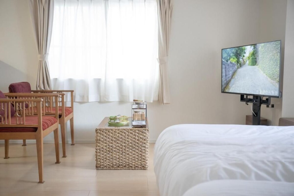A hotel room with a bed, a television, a wooden chair, a small table, and a window with curtains.