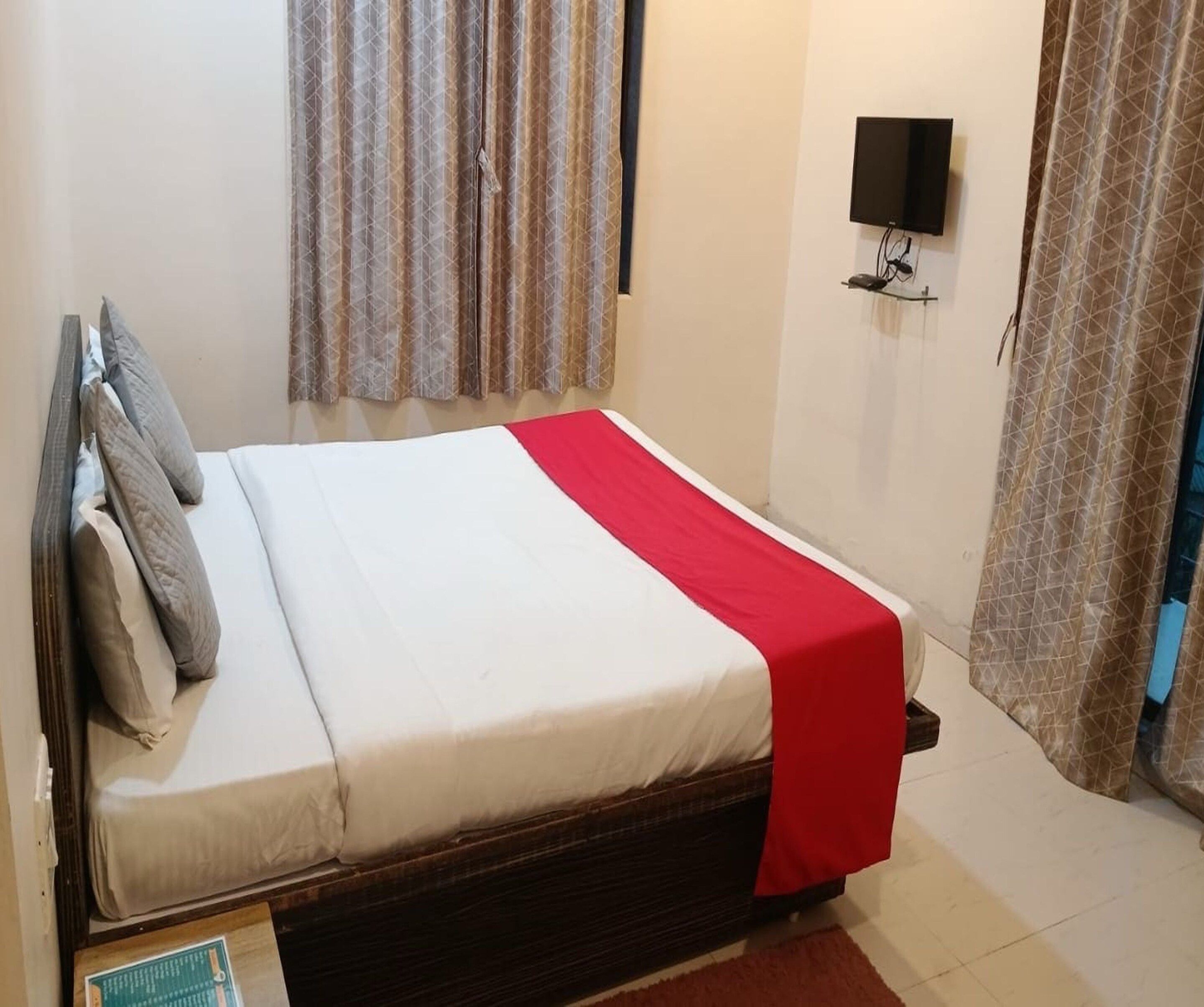 A single bed with a white and red bedspread, a wooden headboard, and a red throw blanket. There is a wall-mounted TV and a window with patterned curtains.