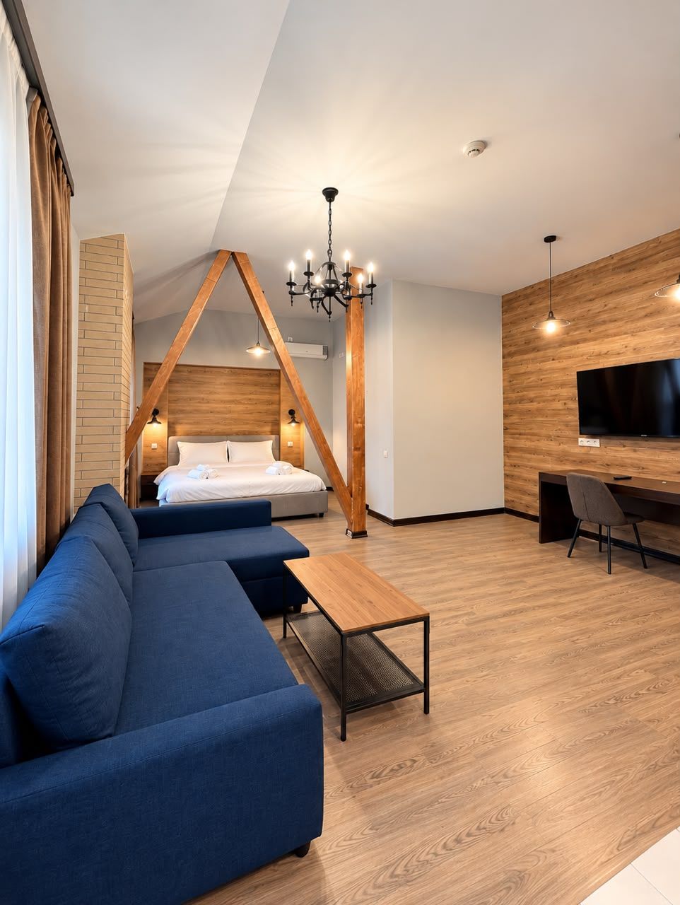 A modern hotel room with a loft bed, a blue sofa, a wooden coffee table, and a TV mounted on the wall.