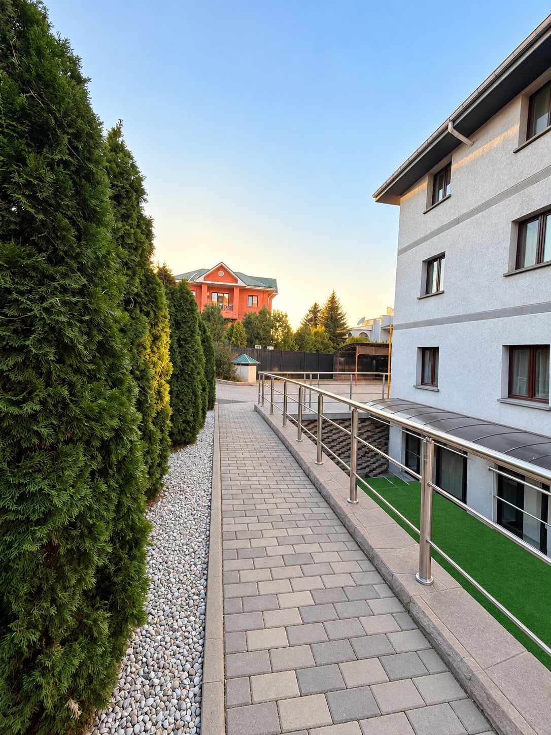 A pathway leading to a building with a red roof, surrounded by greenery and a paved area with a metal railing.
