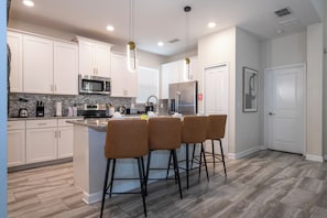 A modern kitchen with white cabinets, a stainless steel refrigerator, a microwave, and a coffee maker. There are three bar stools at the island. - Near Disney 5BR Pool Home with Themed Kids Rooms (Kissimmee)