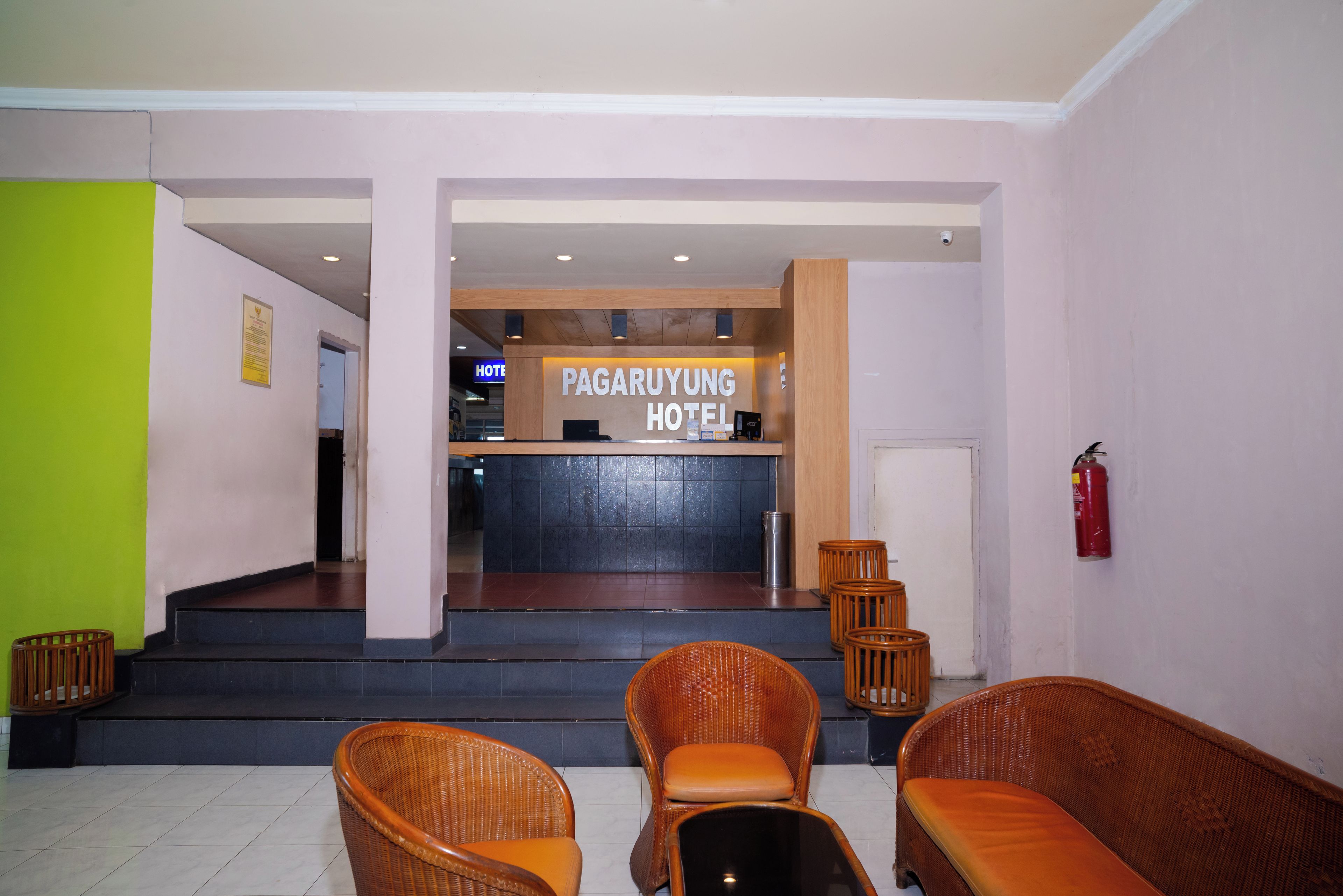 Zona de recepción de un hotel con mostrador, extintor y sillas de mimbre.