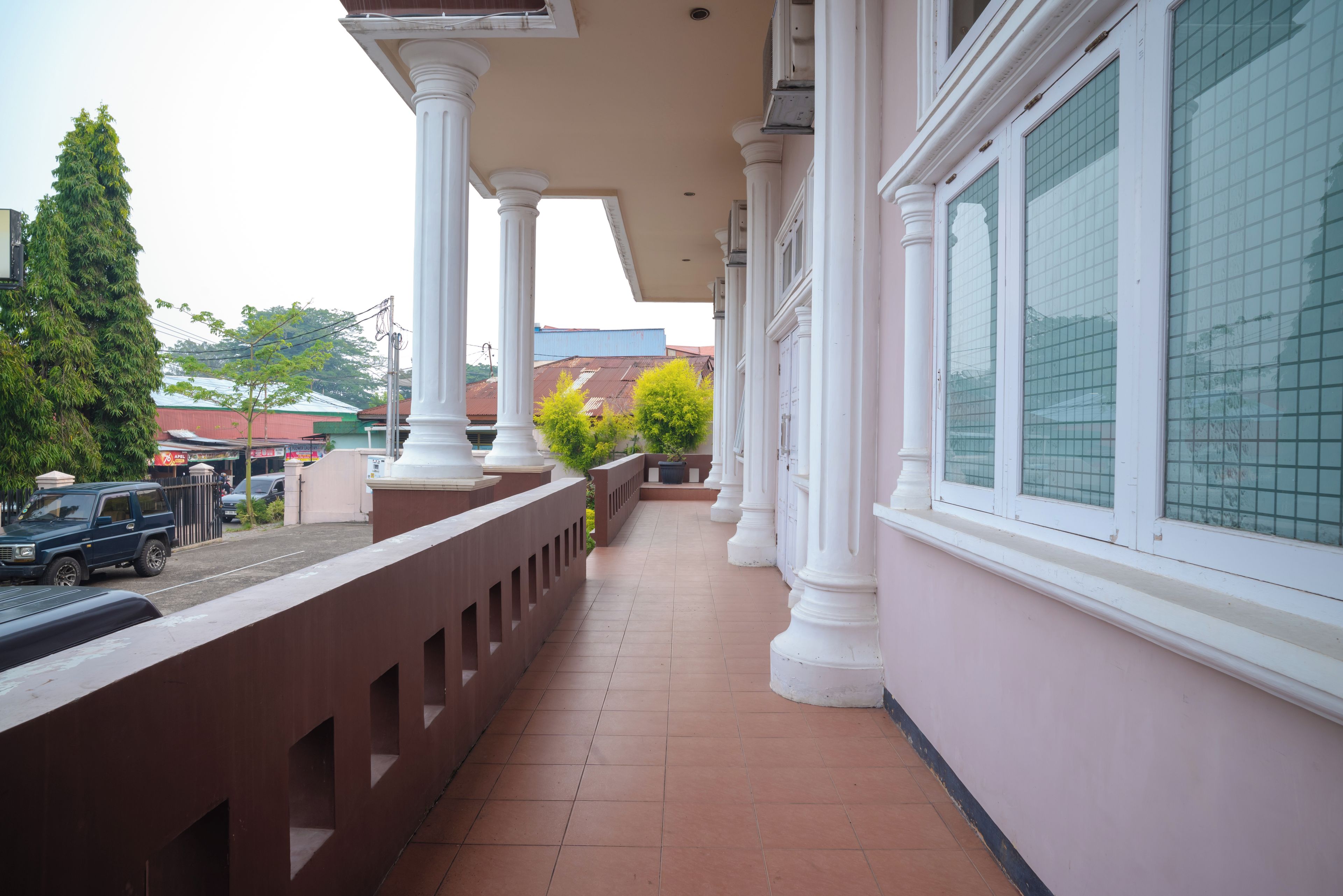 A covered balcony with white columns and a tiled floor, overlooking a street with parked cars and buildings.