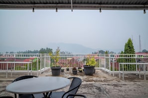 A rooftop terrace with a table and chairs, potted plants, and a view of buildings and mountains in the distance.