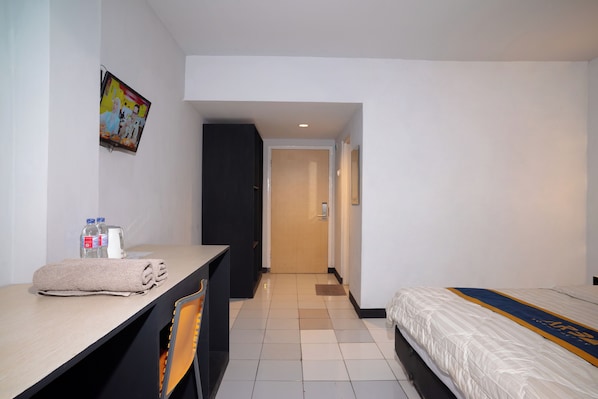 A hotel room with a bed, a desk with a chair, a small table with a towel, a water bottle, a coffee maker, and a painting on the wall.