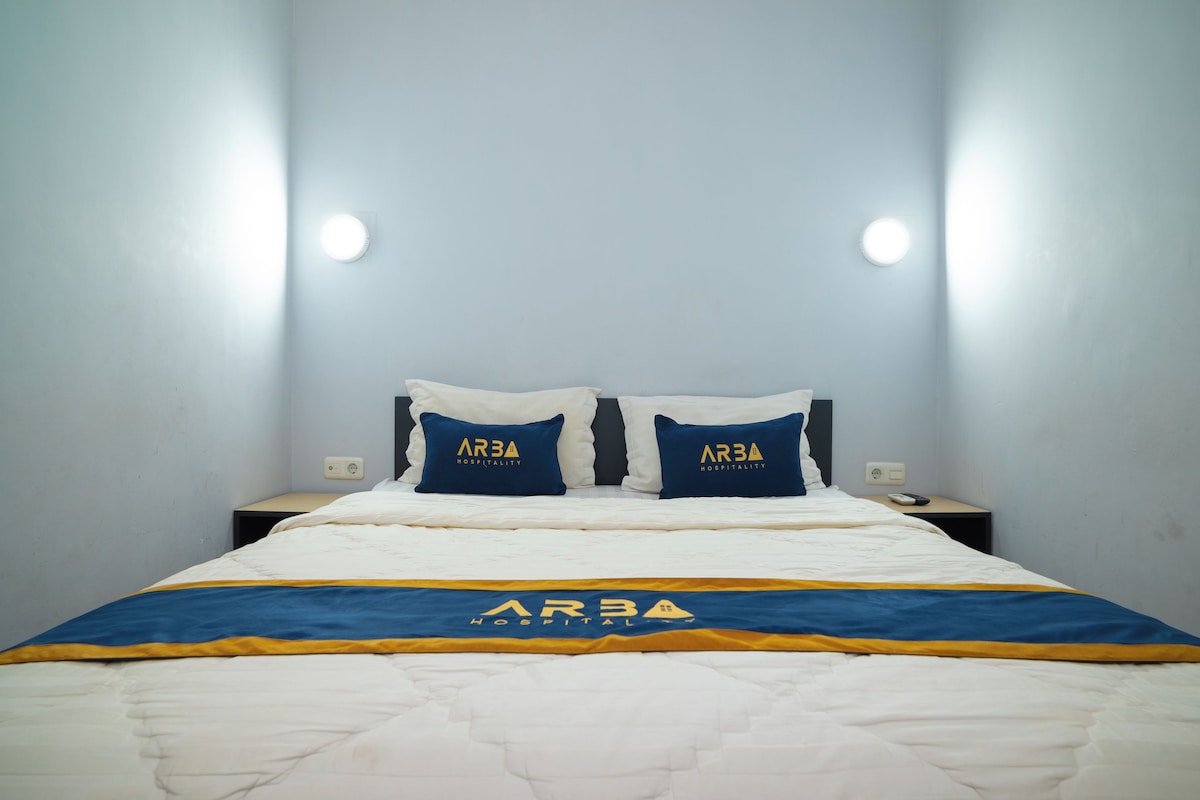 Una habitación de hotel con una cama, dos almohadas con la marca 'ARBA HOSPITALITY', una mesita de noche y una lámpara de pared.
