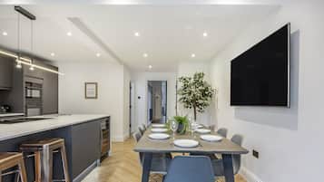 A modern kitchen with a central island, a dining area with a long table set for six, and a flat-screen TV mounted on the wall.