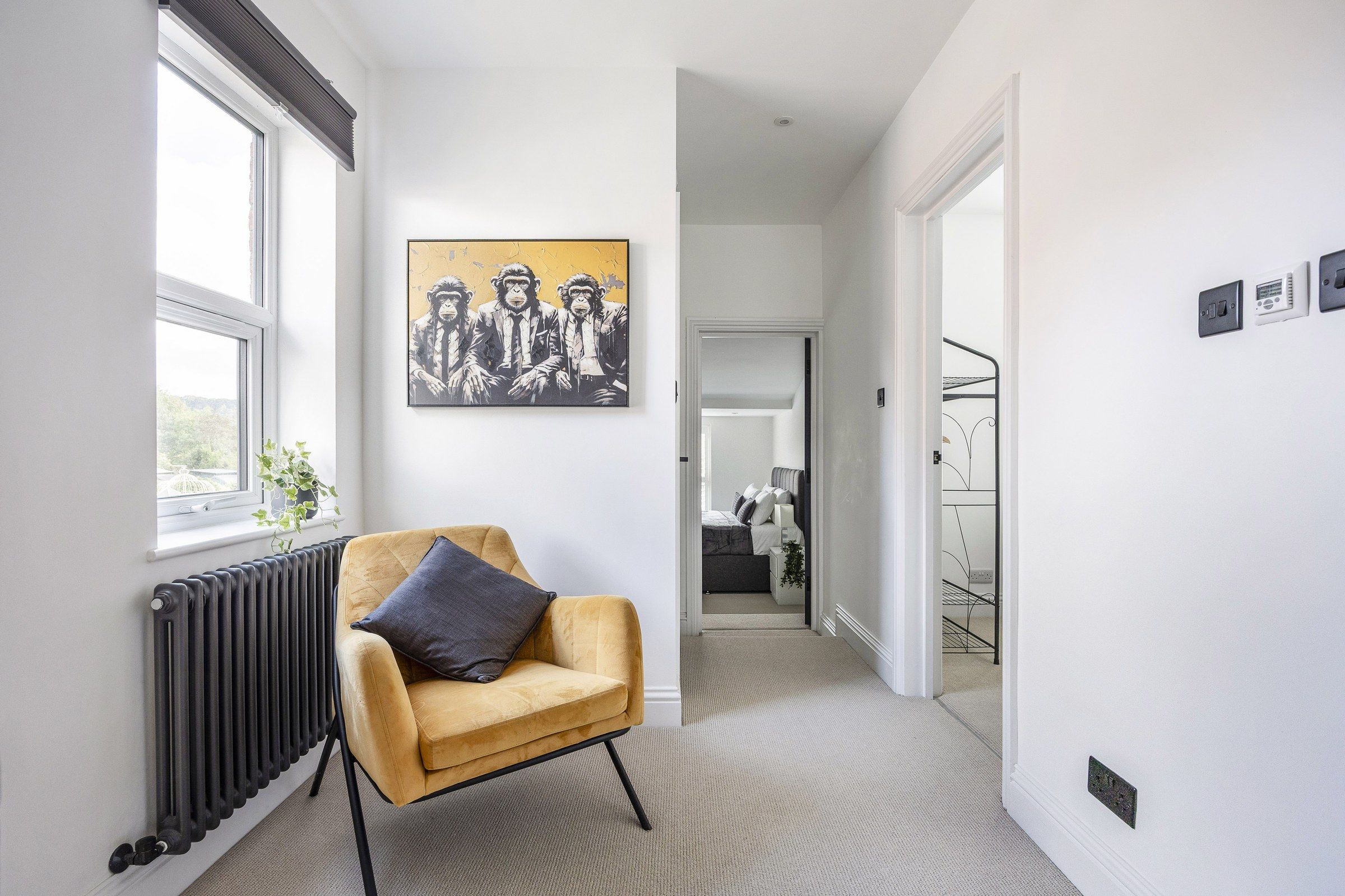 Een gang met een radiator, een gele fauteuil met een kussen, en een schilderij aan de muur.