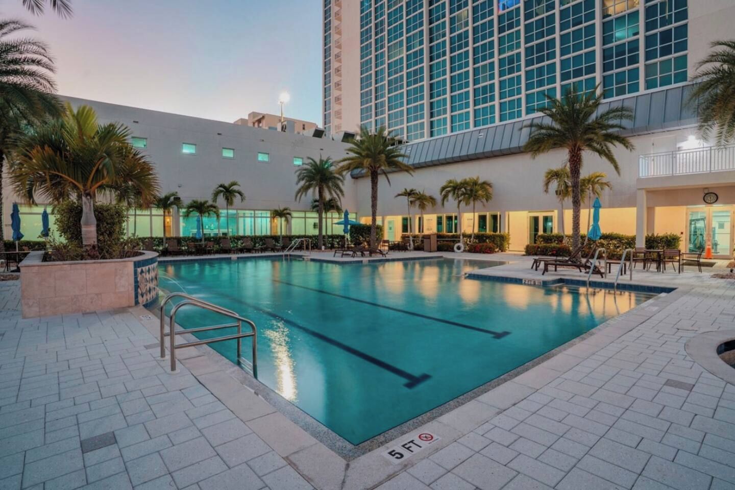 An outdoor swimming pool with a tiled deck, surrounded by palm trees and a modern building in the background.