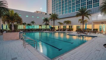 An outdoor swimming pool with a tiled deck, surrounded by palm trees and a modern building in the background.