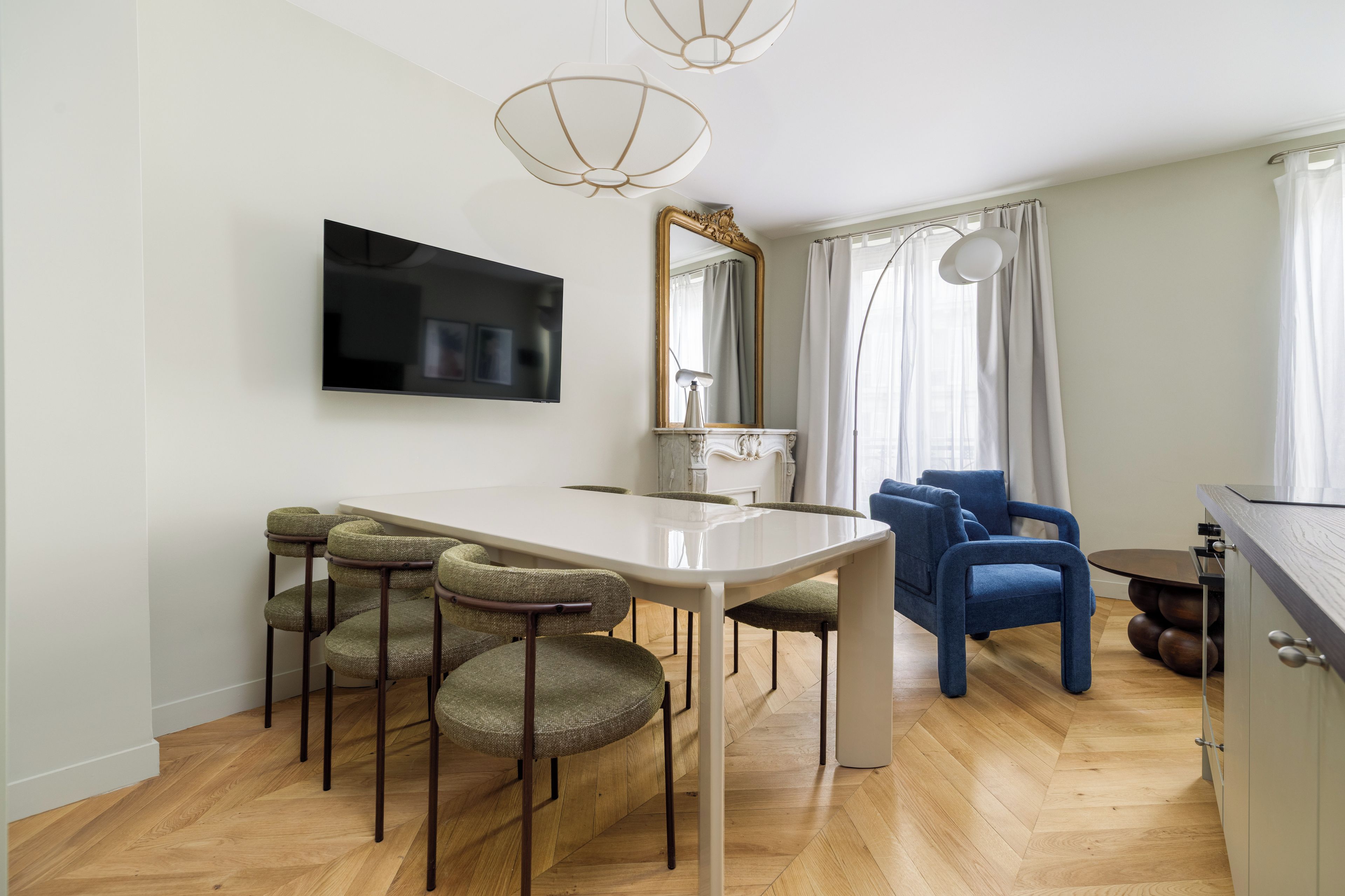Een moderne eetruimte met een witte tafel, vier groene fauteuils, een blauwe stoel en een flatscreen televisie aan de muur.