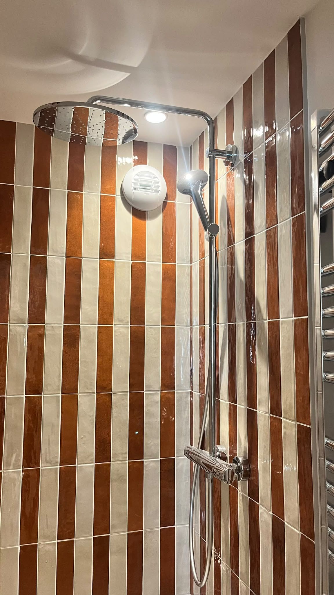 Eine Dusche mit chromfarbenen Armaturen und einer Wand mit vertikal angeordneten braunen und weißen Fliesen.