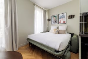 Un dormitorio con cama, una silla, un ventanal con cortinas y dos obras de arte colgadas en la pared.