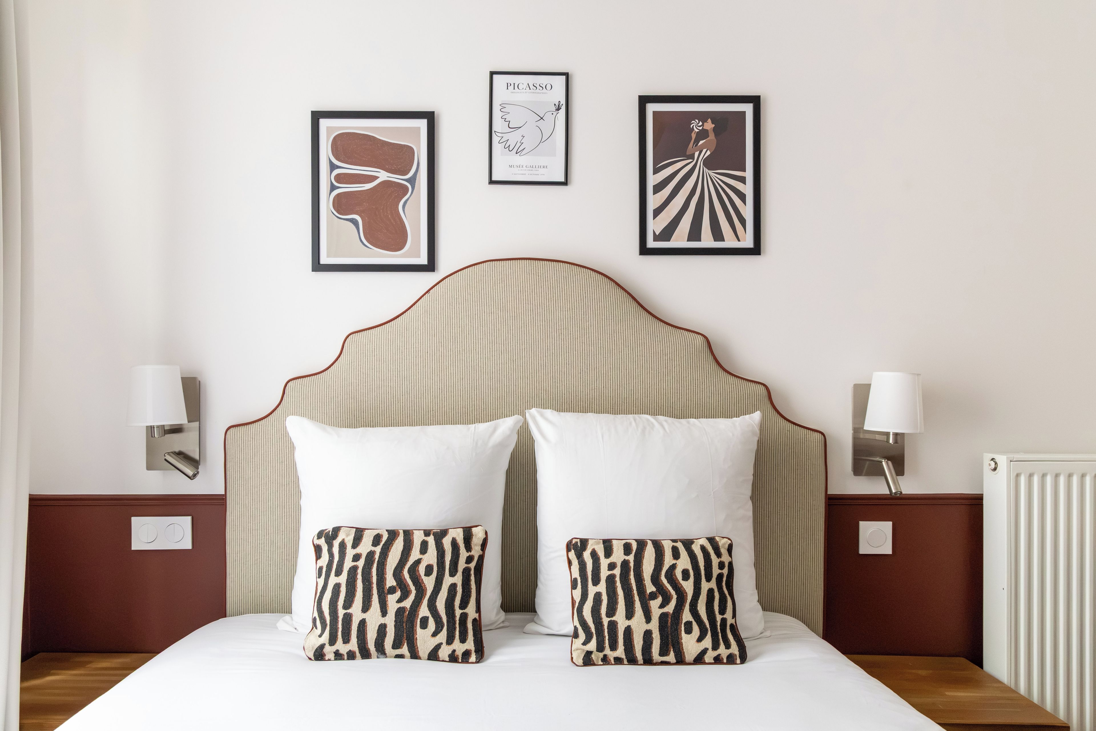 Habitación de hotel con una cama, dos almohadas, un cabecero, dos mesitas de noche y tres cuadros enmarcados en la pared.