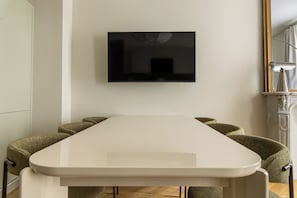 Un comedor moderno con una mesa blanca y sillas tapizadas en verde. Un televisor de pantalla plana está montado en la pared sobre la mesa.