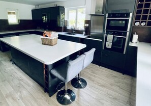 A modern kitchen with a central island, built-in appliances, and a seating area with a chair and bench. - Cornwall Holiday Home with hot tub and acre of garden (Saltash)
