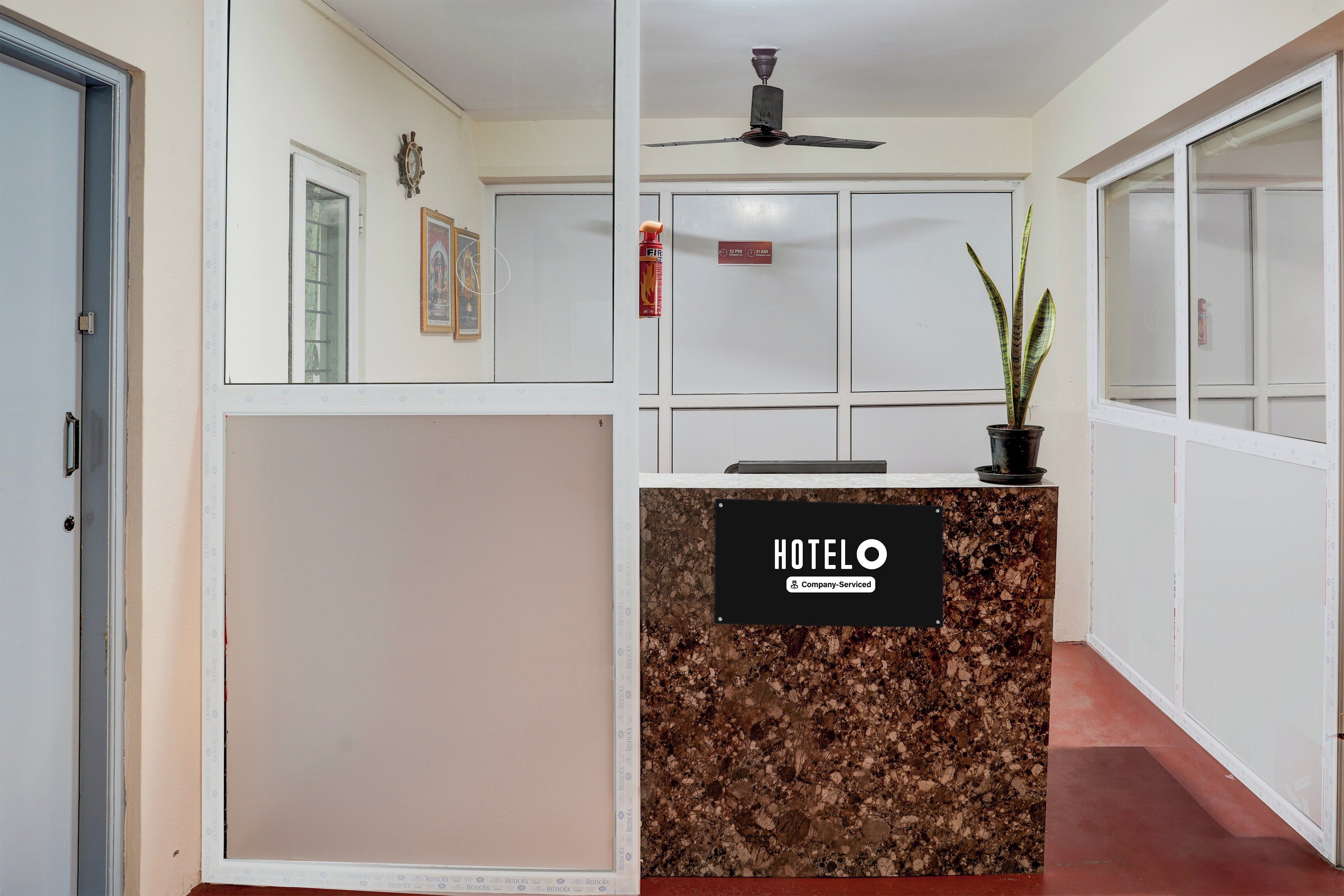 Zona de recepción de un hotel con mostrador, ventilador de techo, cuadros en la pared y extintor.