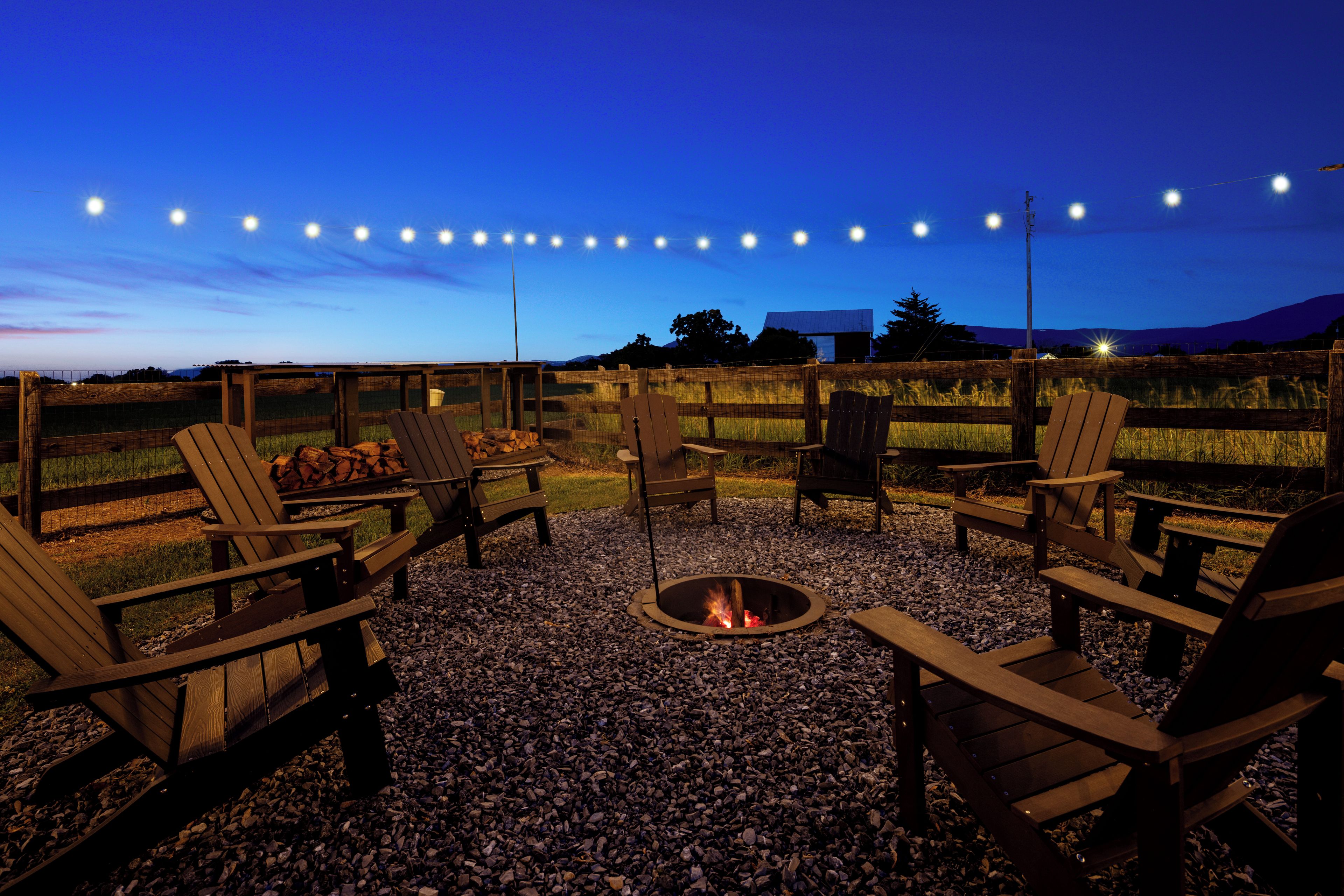 An outdoor seating area with Adirondack chairs and a fire pit, surrounded by a wooden fence and gravel.