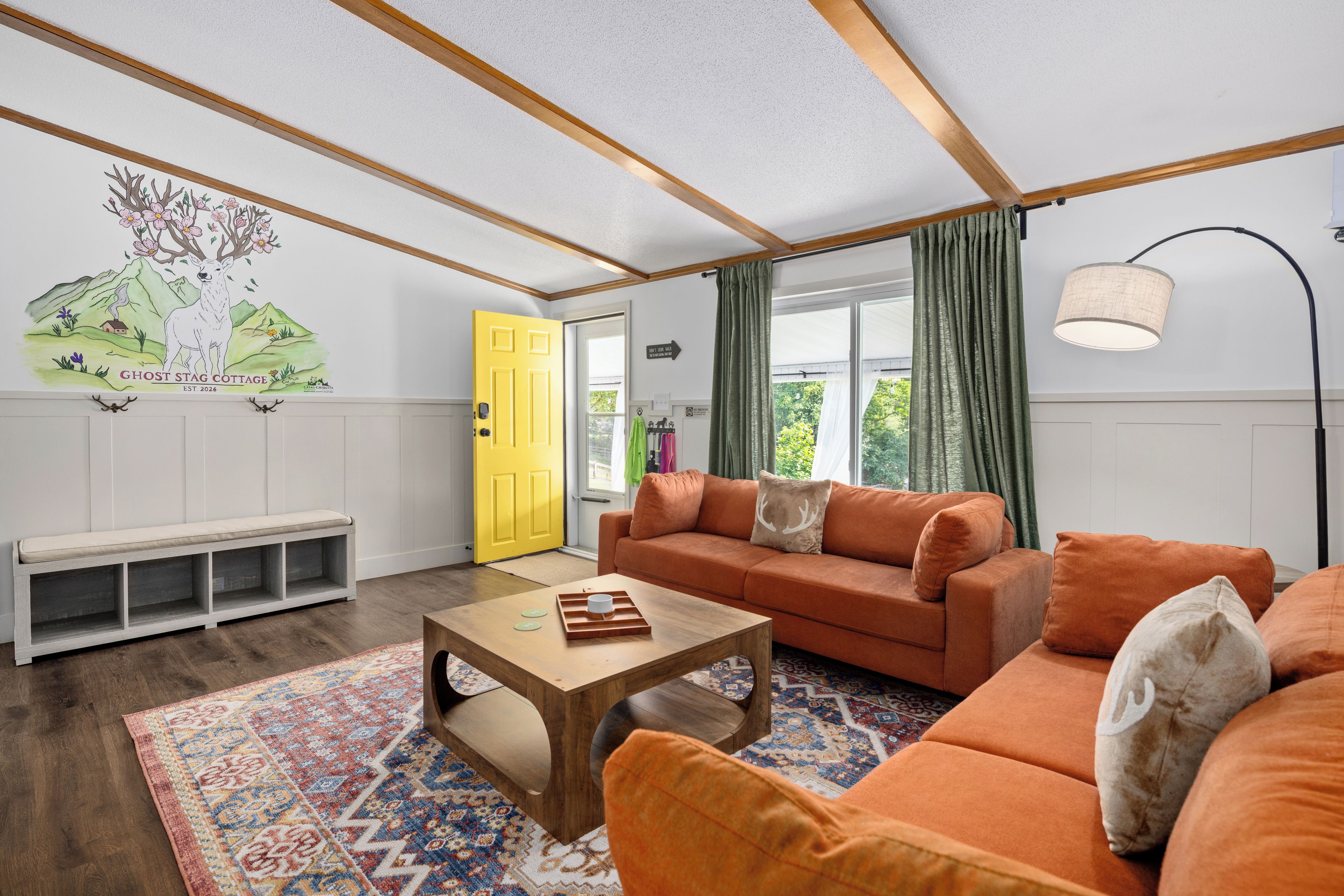 Ein modernes Wohnzimmer mit einer Couch, einem Couchtisch und einer Bank. Eine gelbe Tür und ein Wandgemälde sind zu sehen.