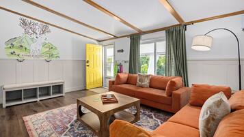 Ein modernes Wohnzimmer mit einer Couch, einem Couchtisch und einer Bank. Eine gelbe Tür und ein Wandgemälde sind zu sehen.