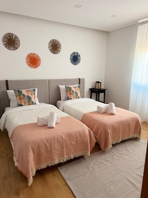 Ein Doppelzimmer mit zwei Betten, jeweils mit einer gestreiften Decke und einem Kissen. Über den Betten befinden sich Wandbilder und neben dem Fenster ein kleiner Tisch mit Büchern.