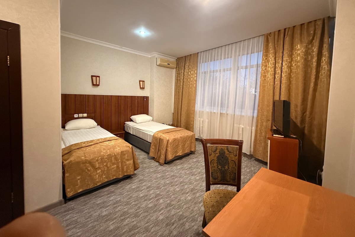 Een hotelkamer met twee bedden, een tafeltje, een stoel, een televisie en een raam met gordijnen.