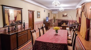 Een statige eetzaal met een lange tafel, stoelen, een kroonluchter en een televisietoestel.