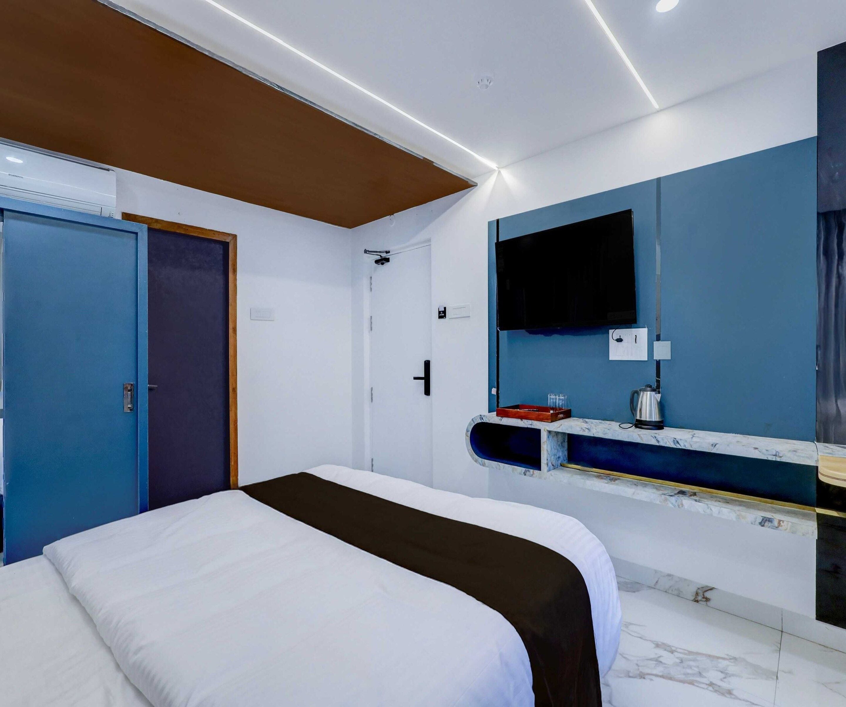 A hotel room with a bed, a television mounted on the wall, and a small shelf with a kettle and other items.