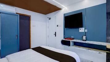 A hotel room with a bed, a television mounted on the wall, and a small shelf with a kettle and other items.