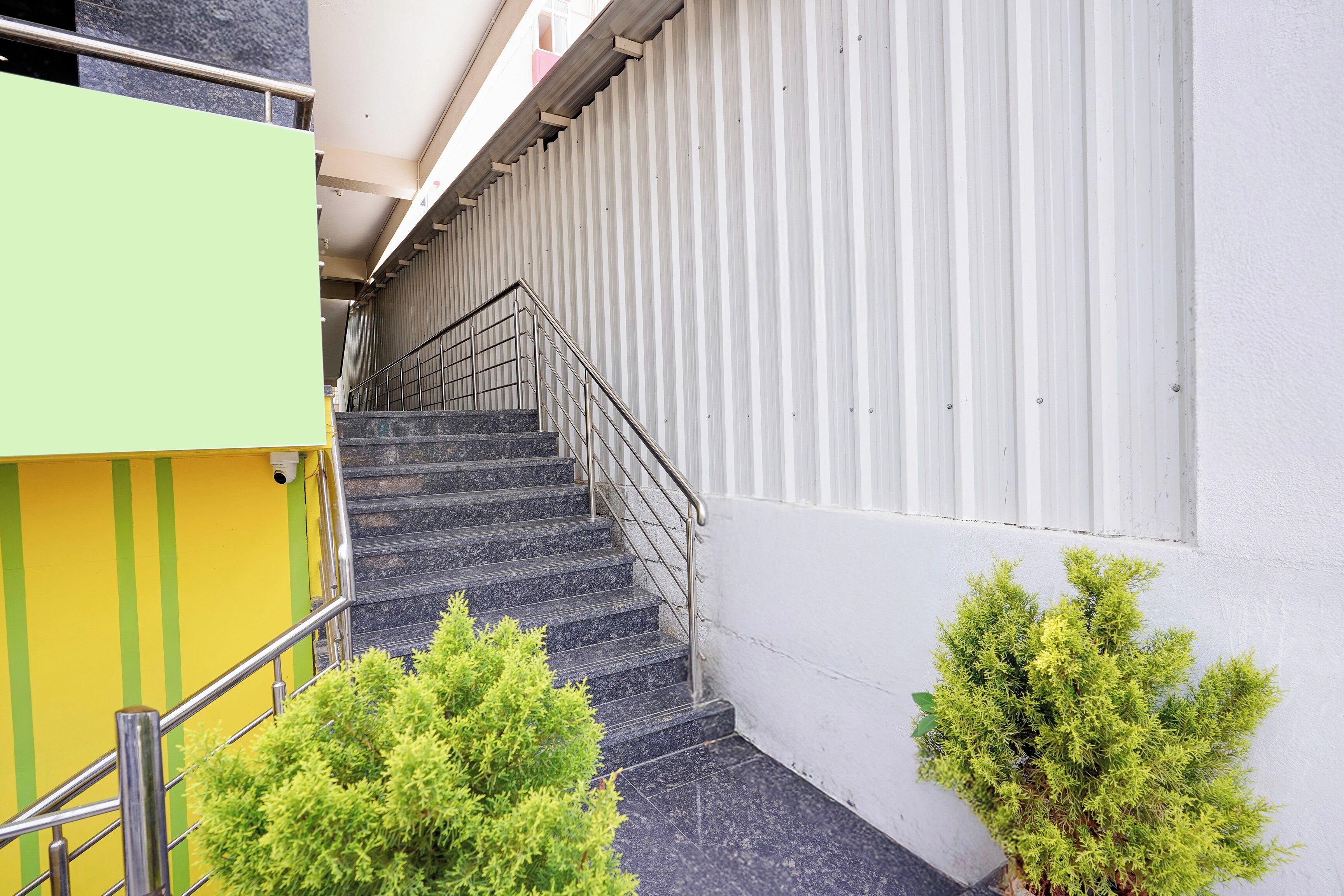 Una escalera con barandillas de metal que sube a un piso superior, flanqueada por un edificio verde y amarillo y una pared blanca con líneas verticales.