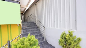 A staircase with metal railings leading to an upper floor, flanked by a green and yellow building and a white wall with vertical lines.