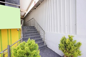 Una escalera con barandillas de metal que sube a un piso superior, flanqueada por un edificio verde y amarillo y una pared blanca con líneas verticales.