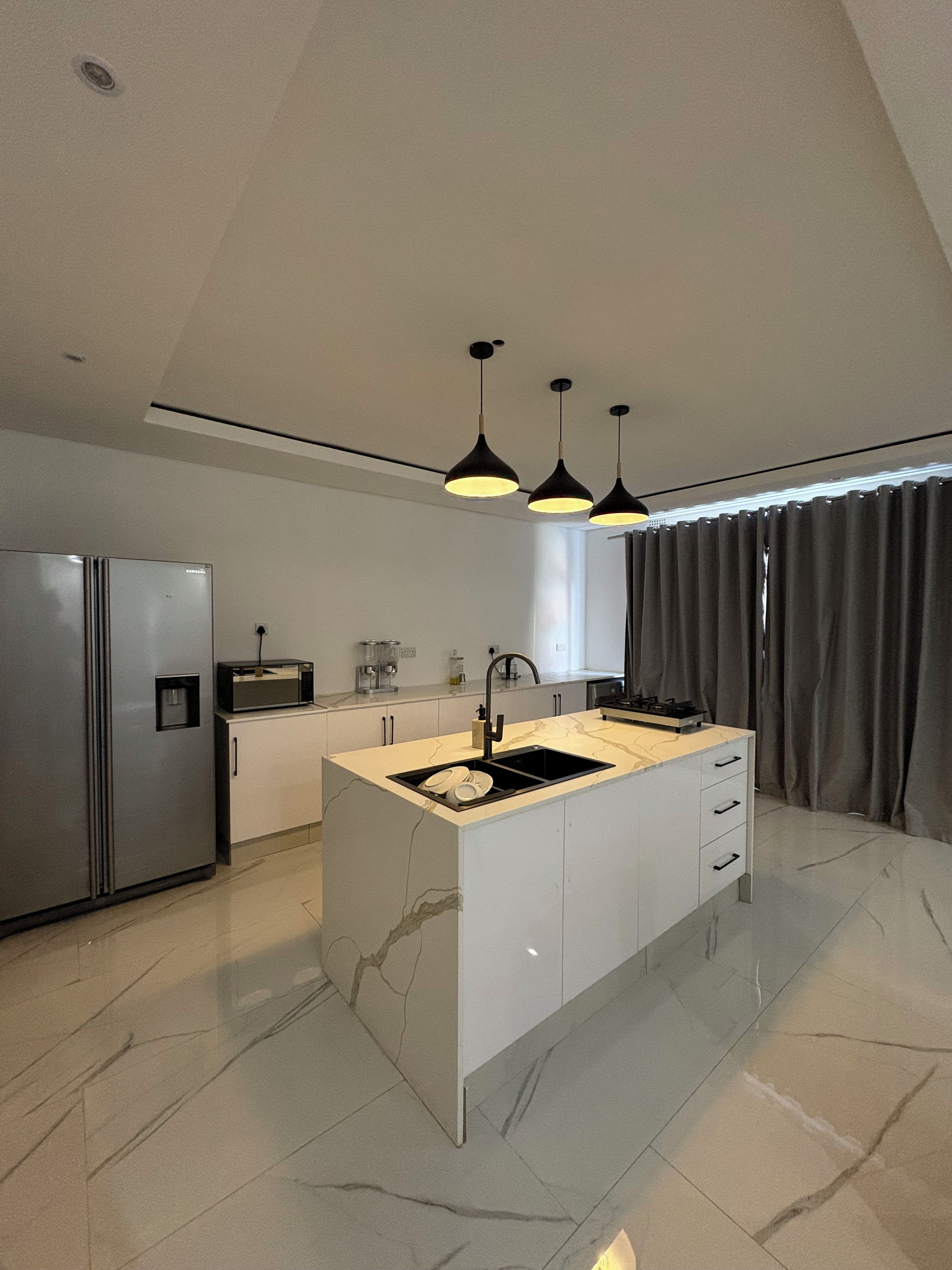 A modern kitchen with a central island, stainless steel appliances, pendant lighting, and a large window with curtains.