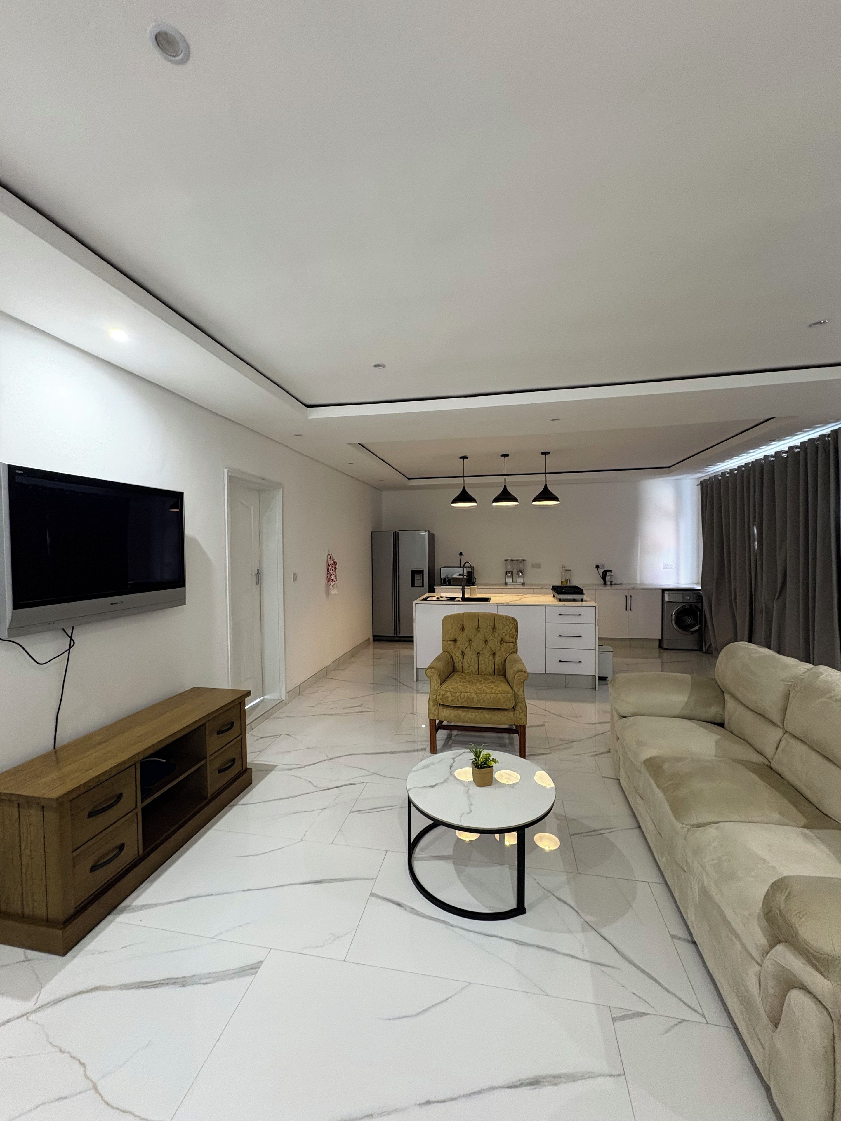 A modern living room with a flat-screen TV, a sofa, a coffee table, and a kitchen area in the background.