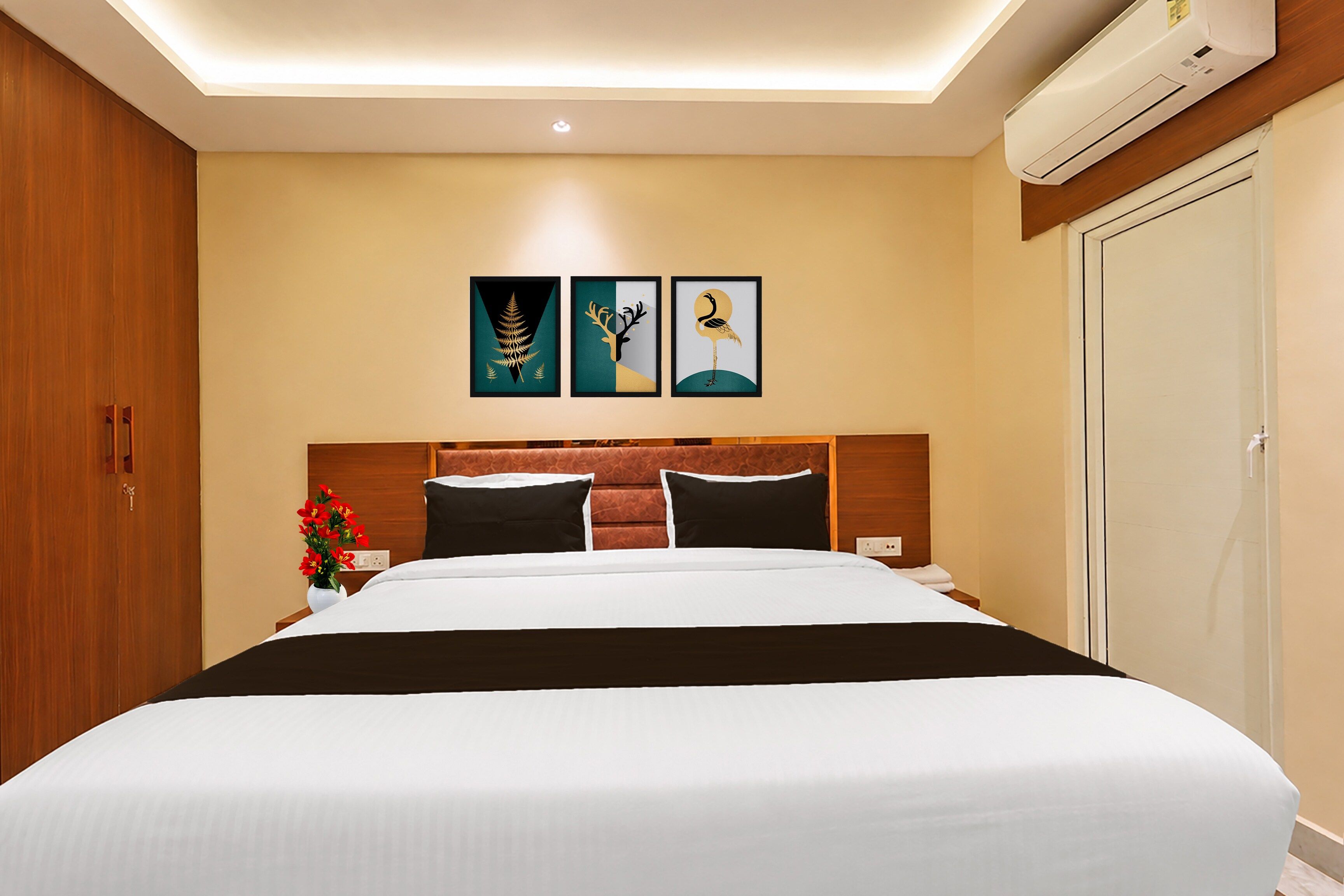 A hotel room with a bed, a nightstand, a vase with flowers, and three framed artworks on the wall.