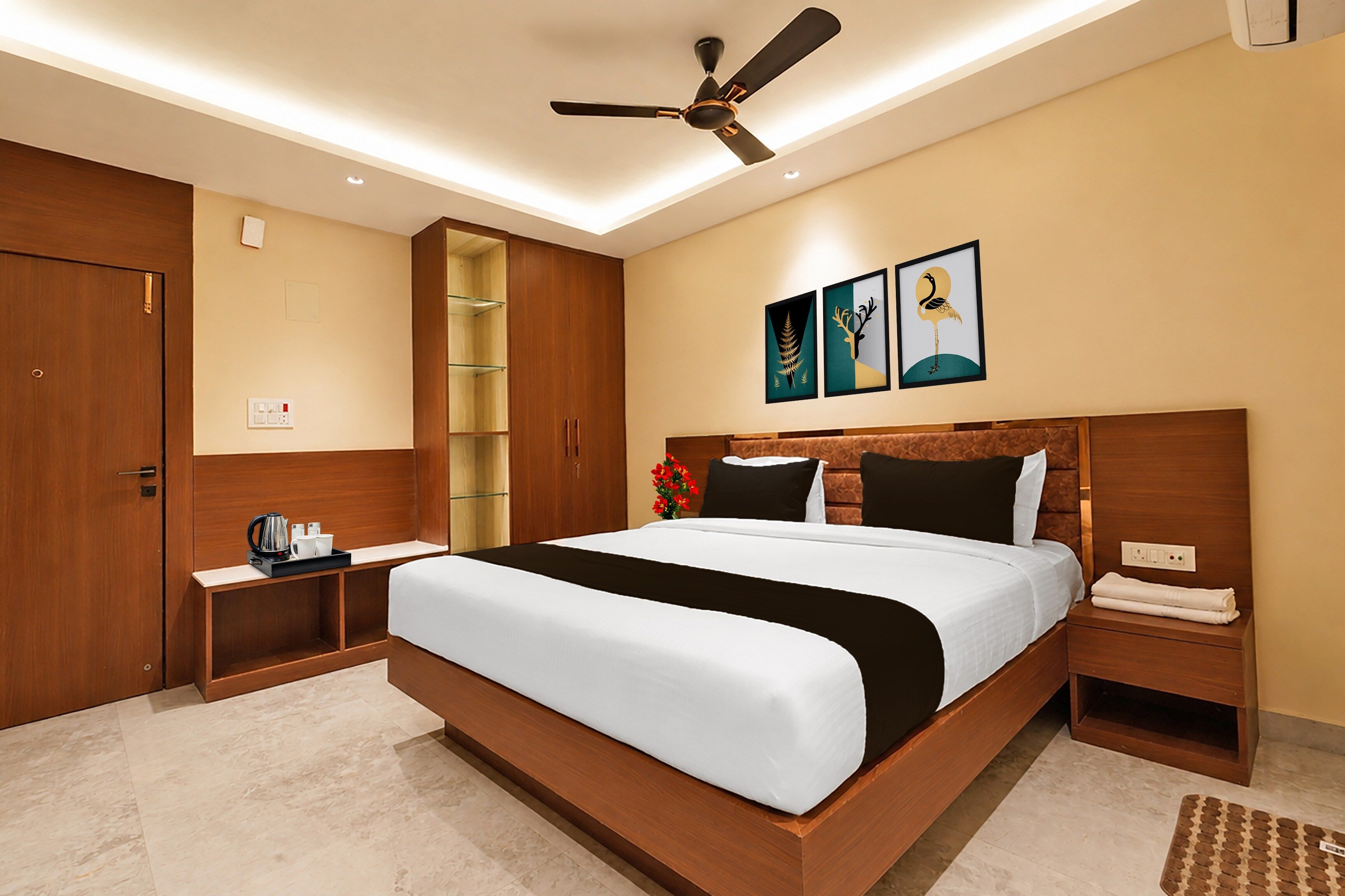 Una habitación de hotel con una cama grande, un ventilador de techo, una mesita de noche con cafetera y obras de arte en la pared.
