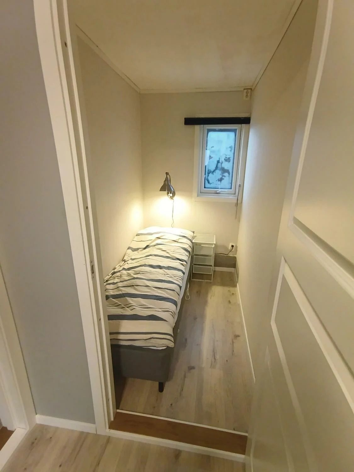 Een kleine kamer met een bed, nachtkastje, lamp en raam.