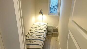 Een kleine kamer met een bed, nachtkastje, lamp en raam.