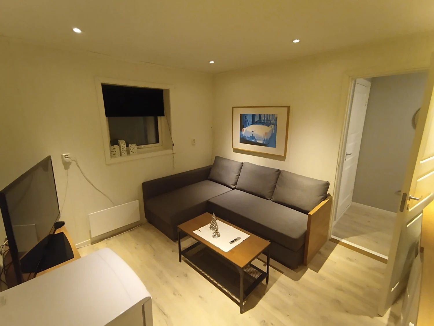 Een woonkamer met een bank, salontafel, televisie, raam met zonwering en een schilderij aan de muur.