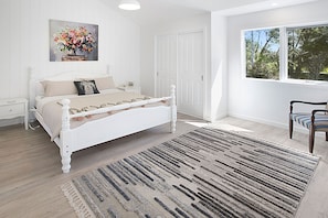 Ein Schlafzimmer mit einem weißen Bett, einem Nachttisch, einem Sessel, einem Fenster mit Blick auf Bäume und einem Bild an der Wand.