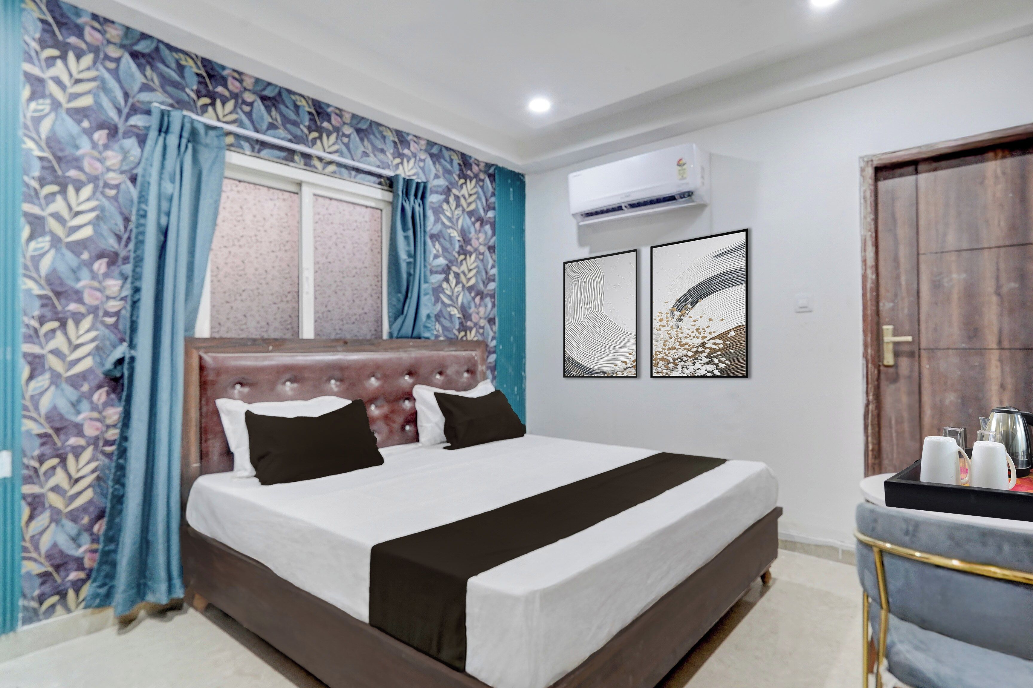 A hotel room with a bed, curtains, a window, wall art, an air conditioning unit, and a door.