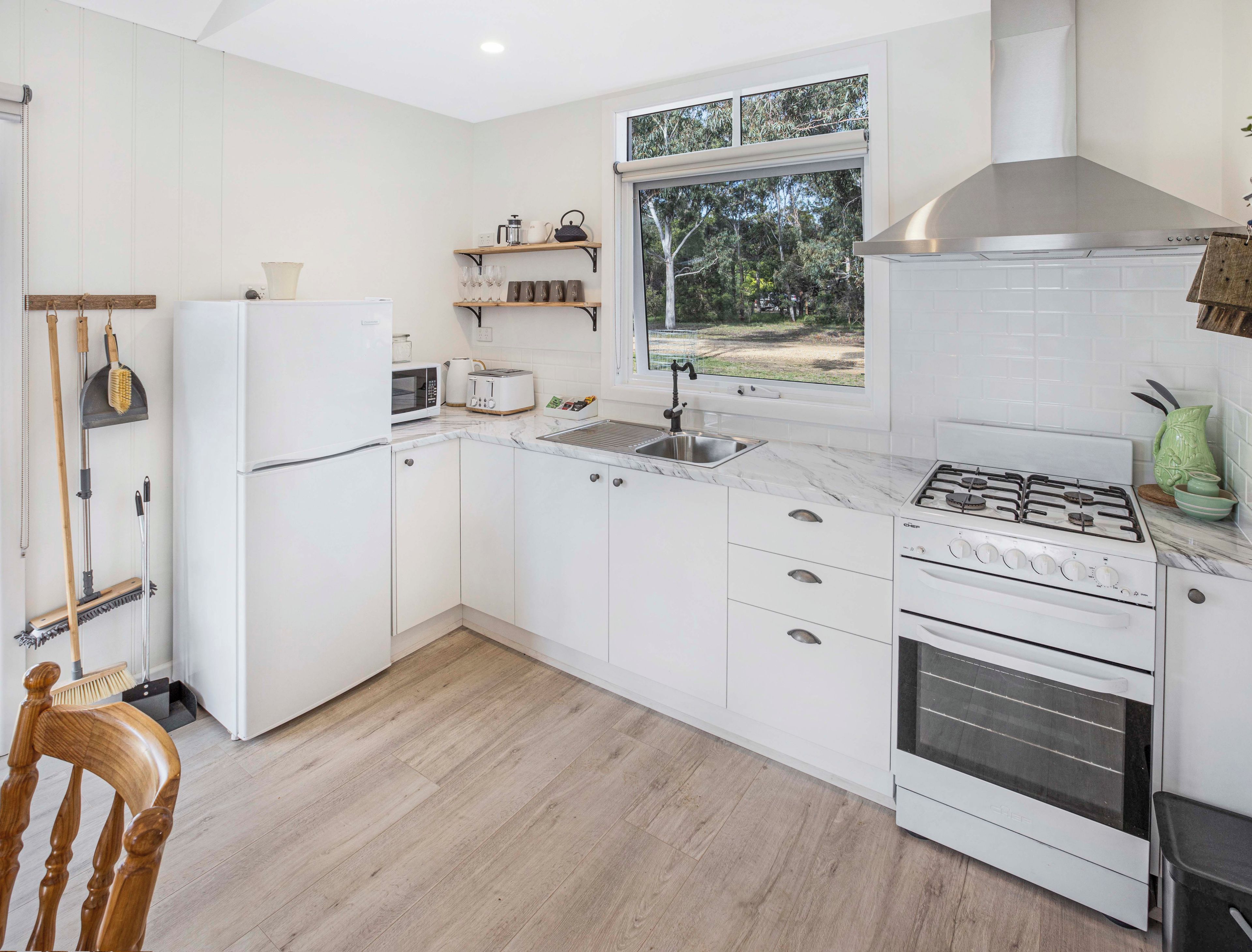 Una cocina moderna con muebles blancos, un lavamanos de acero inoxidable, horno empotrado y ventana con vistas a un jardín.