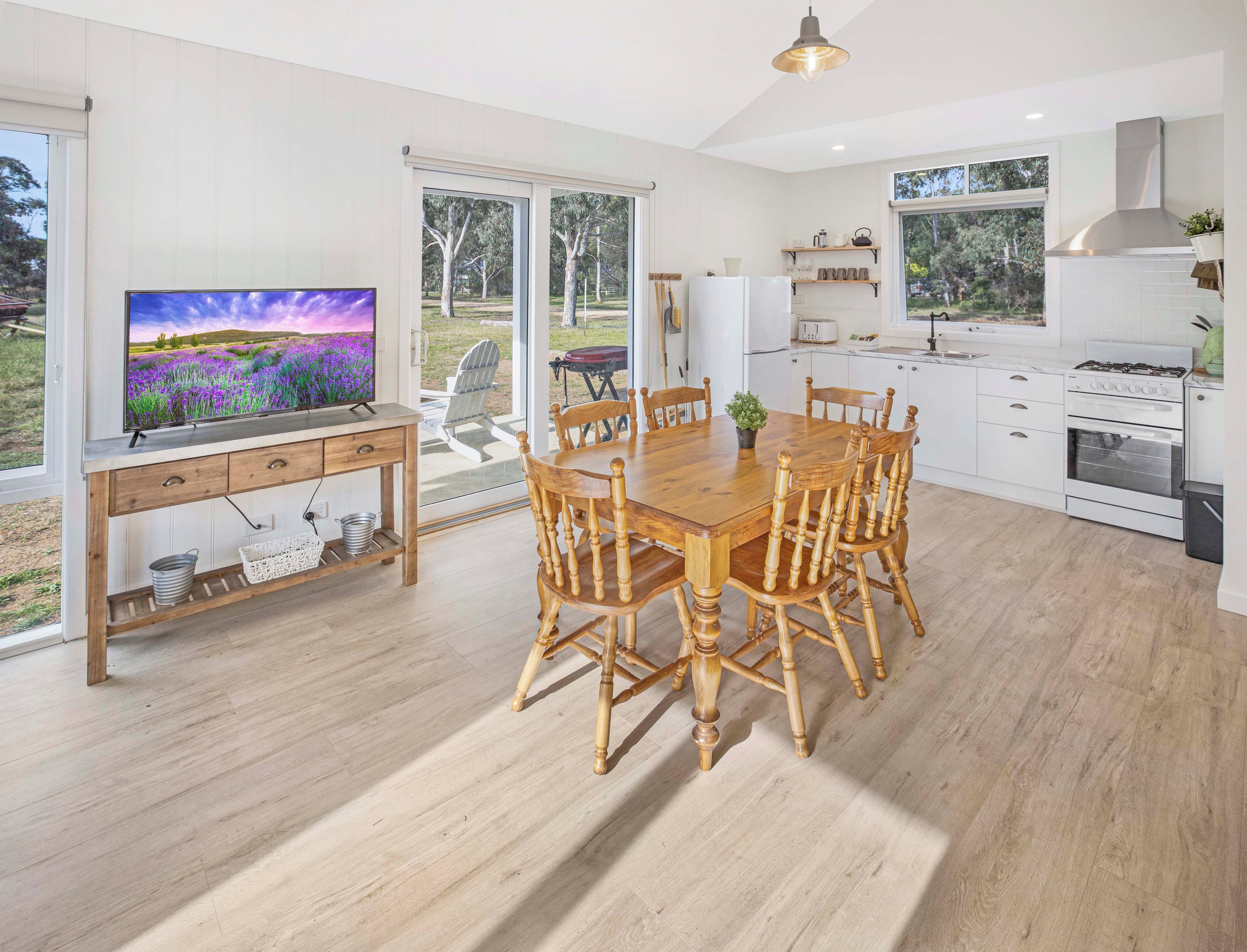 A modern kitchen with white cabinetry, a dining area with wooden chairs, and a television displaying a field of purple flowers.