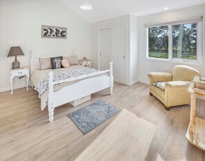 Een slaapkamer met een wit bed, een nachtkastje, een lamp, een stoel en een raam met uitzicht op bomen.