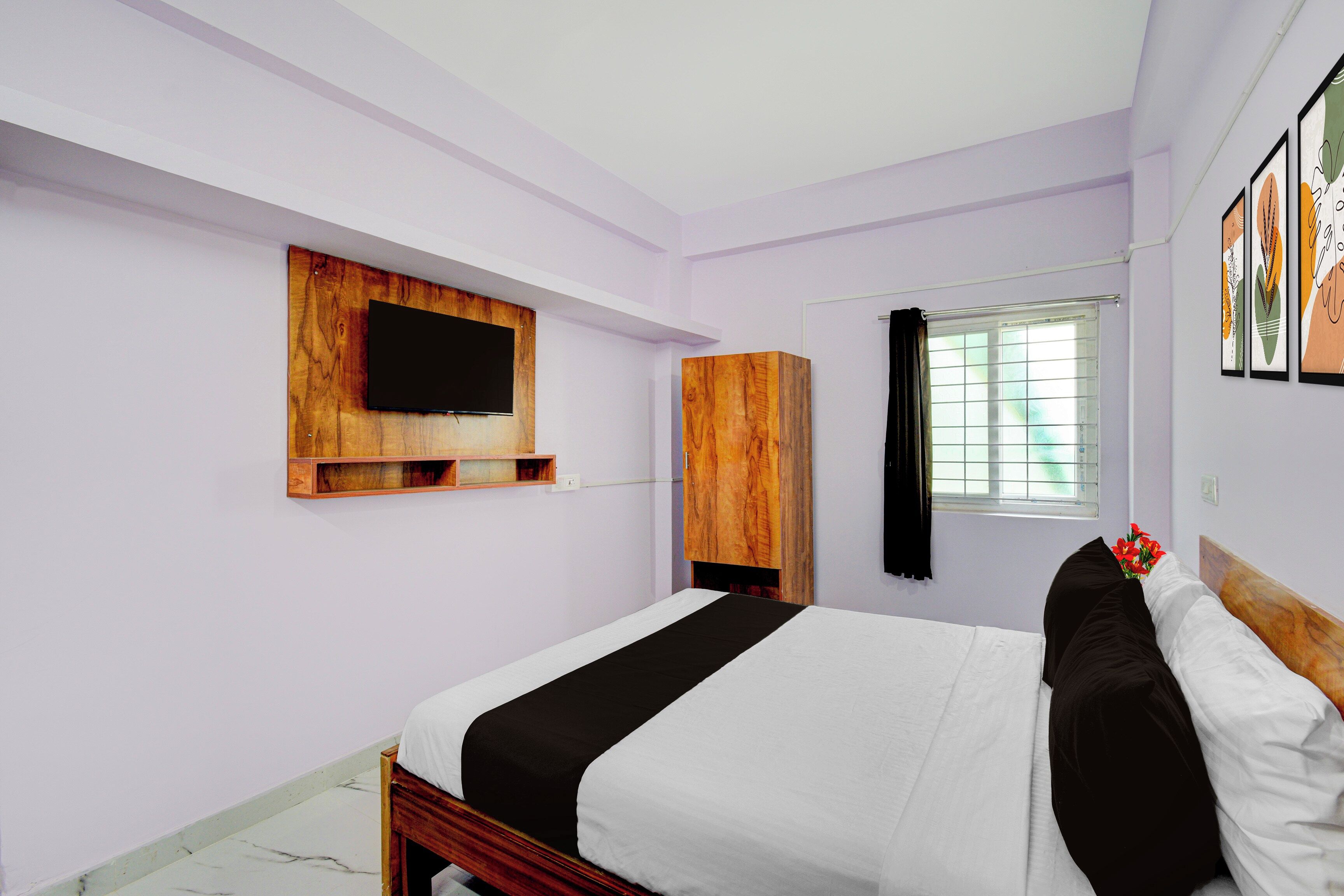 Un dormitorio con cama, televisor montado en un panel de madera, ventana con cortinas y estante fijado a la pared.