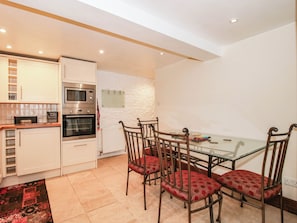 A compact kitchen with modern appliances, a dining area with a glass table and metal chairs, and a patterned rug on the floor. - Stone Cottage (Church Stretton)