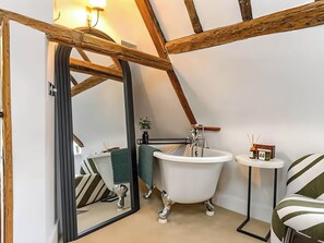 A bathroom with a freestanding bathtub, a mirror, a small table with a candle, and a striped chair. - Jack's Place at Cobnut Cottage (Sandwich)