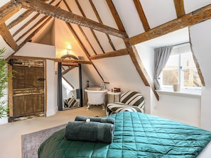 Ein Schlafzimmer mit schrägem Dach, einem großen Bett mit türkisfarbener Steudecke, einer Holztür, einem kleinen Badezimmer mit Waschbecken und Spiegel sowie einem Fenster mit Vorhängen.