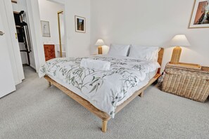 Quarto com cama, duas luminárias de apoio, um cesto de vime e porta que dá para outro cômodo.